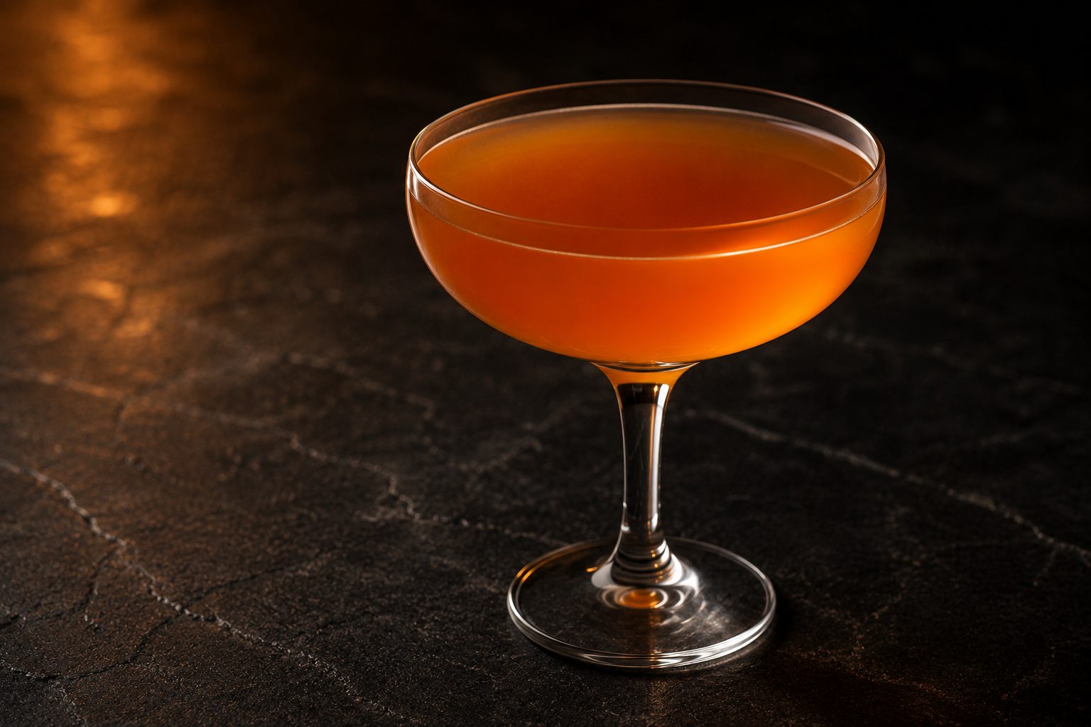A Paper Plane served up in a coupe, deep amber-orange, with no garnish, on a dark stone surface.
